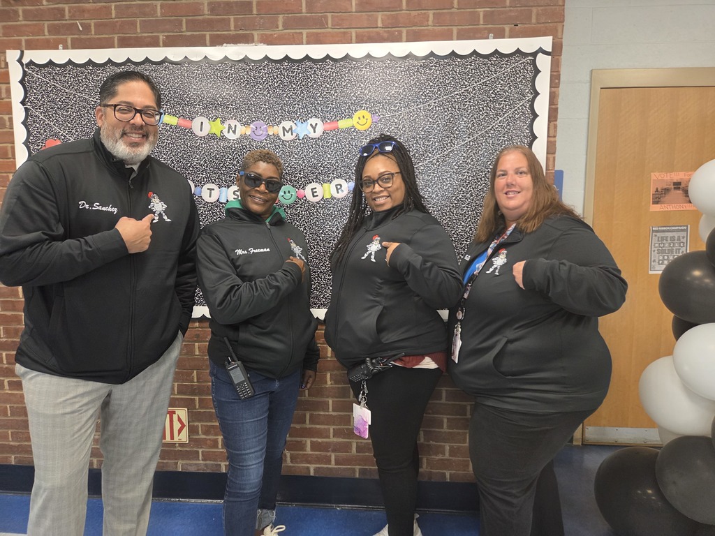Four Southeast Delco School District staff members stand smiling in front of a decorated bulletin board with a black-and-white background and colorful letters that read “IN MY TEACHER ERA.” Each person points to the district logo on their matching black jackets, which feature embroidered names. A black-and-white balloon column decorates the doorway beside them.