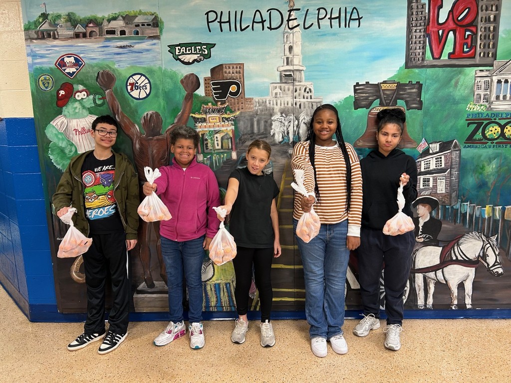 Five students stand smiling in front of a colorful mural featuring Philadelphia landmarks such as the Liberty Bell, the LOVE sculpture, and sports team logos. Each student holds a bag of oranges, proudly displaying them toward the camera.