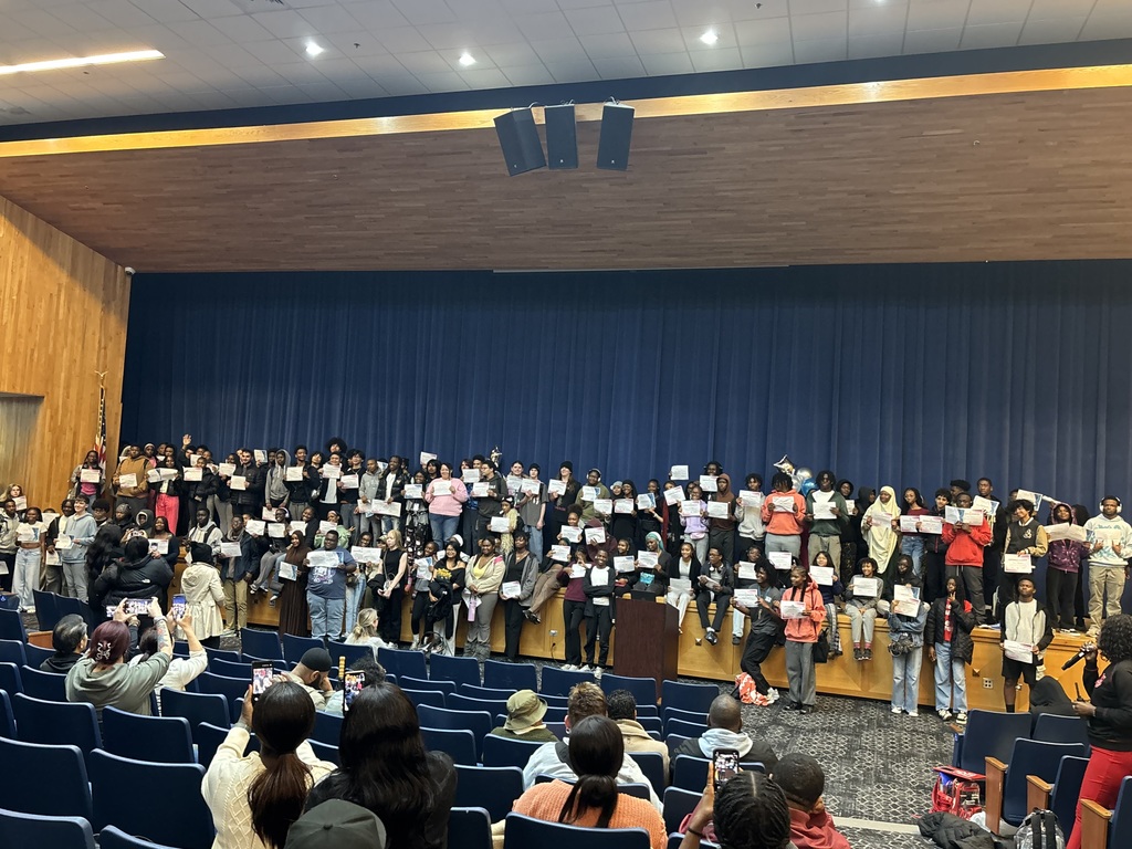 A large group of students stand across a stage holding certificates during the Keystone Award Ceremony. Dozens of family members and guests in the audience take photos from their seats.