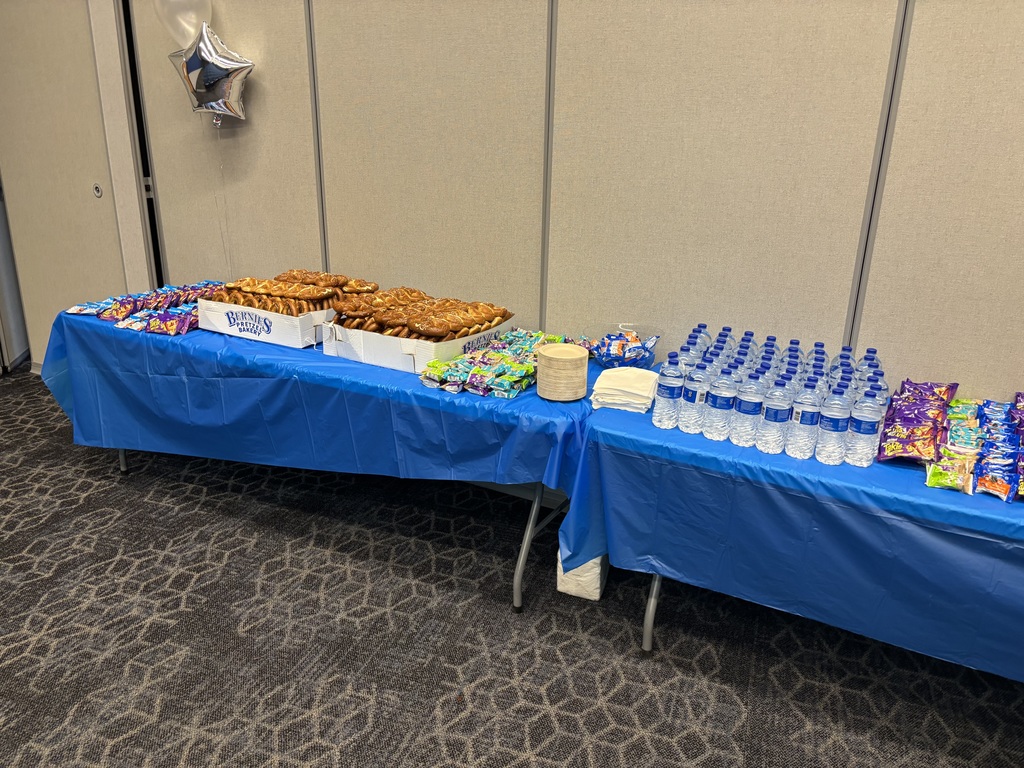 A refreshment table covered with a blue tablecloth is set with trays of pretzels, assorted snacks, water bottles, and napkins.