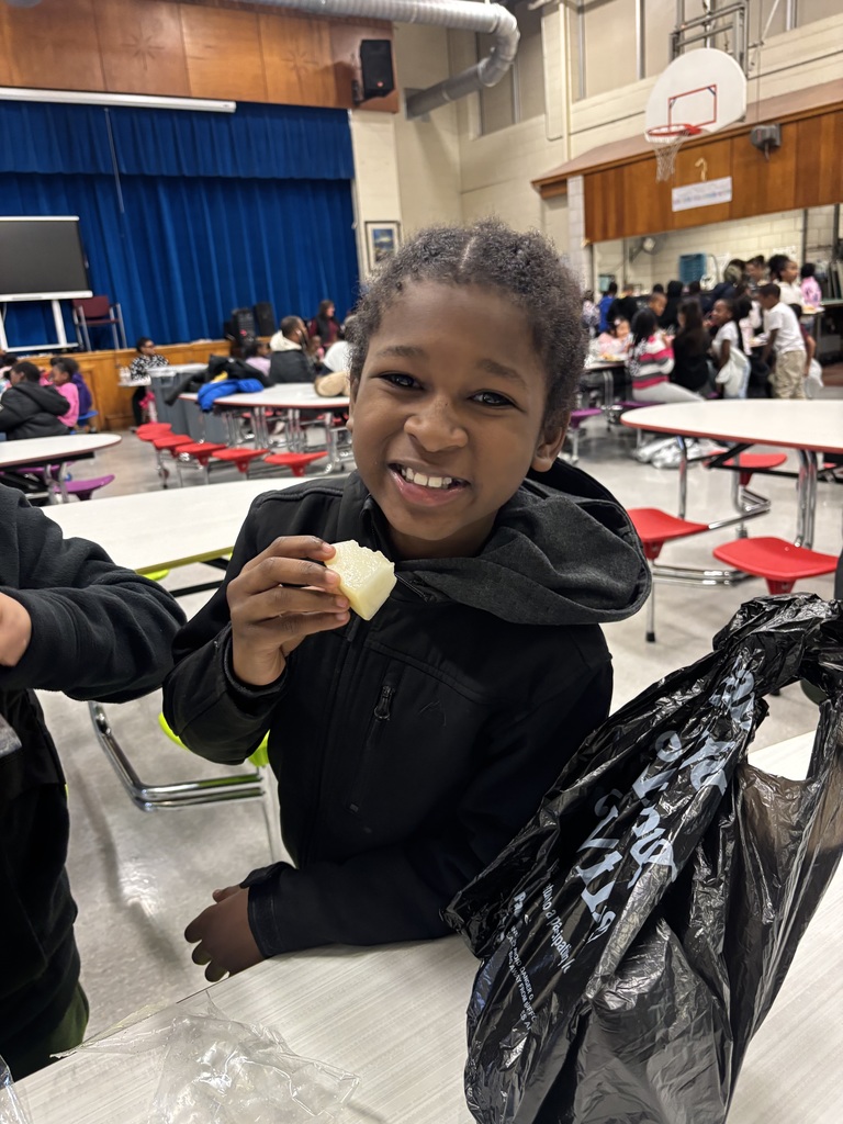 A student smiles at the camera while holding a piece of fruit in a cafeteria.