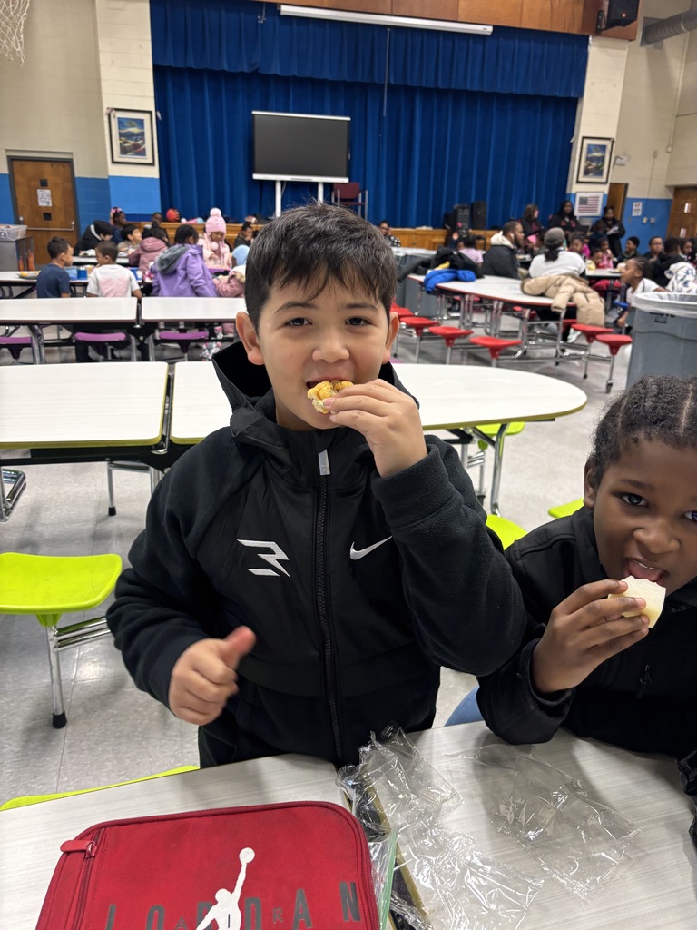 A student holds up a piece of food and gives a thumbs-up while smiling at the camera in a cafeteria.