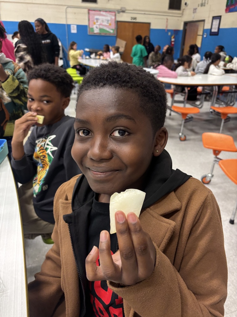 A smiling student sits at a cafeteria table holding a piece of sliced fruit toward the camera.