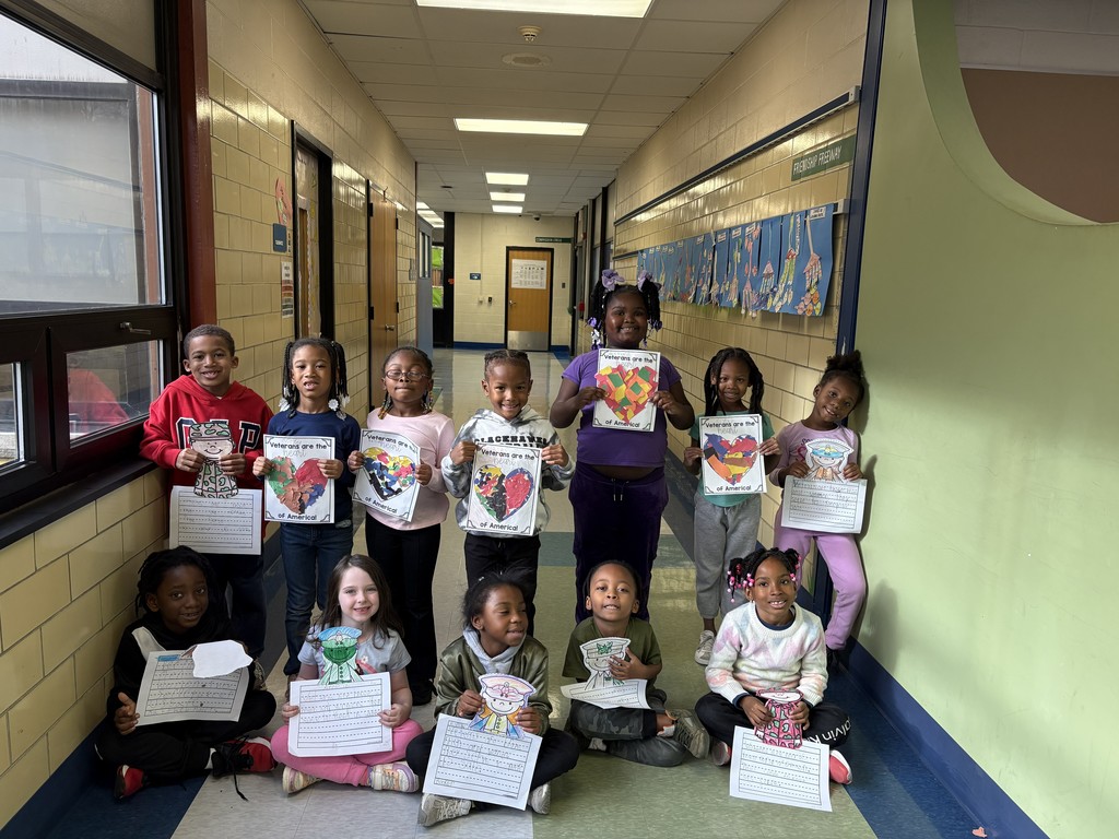 A group of young elementary students pose in a school hallway, smiling and holding up their Veterans Day drawings and writing assignments. Some students stand while others sit on the floor in front. Their artwork features colorful hearts, flags, and messages thanking veterans.