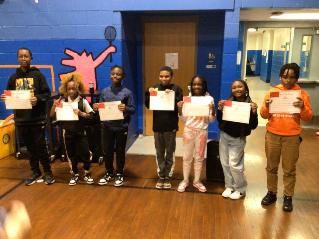 Seven elementary students stand in a gymnasium holding certificates and smiling. They are lined up side by side in front of a blue wall with a painted pink figure holding a badminton racket.