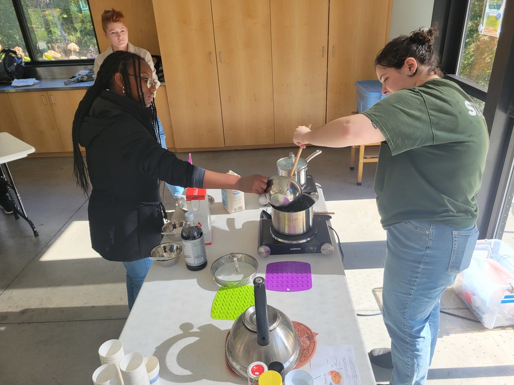 A student pours liquid from a small pot into another container while an instructor stirs a pot on a portable burner.