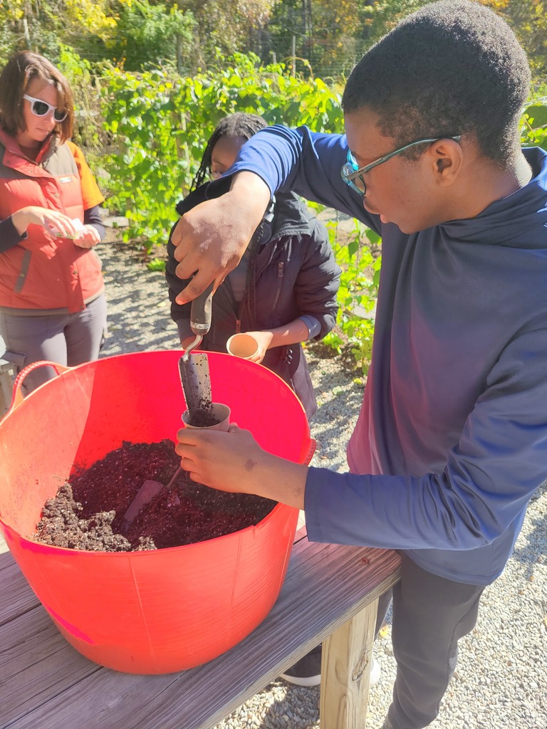 A student scoops soil from a large red bucket into a small paper pot while another student and an adult instructor watch.