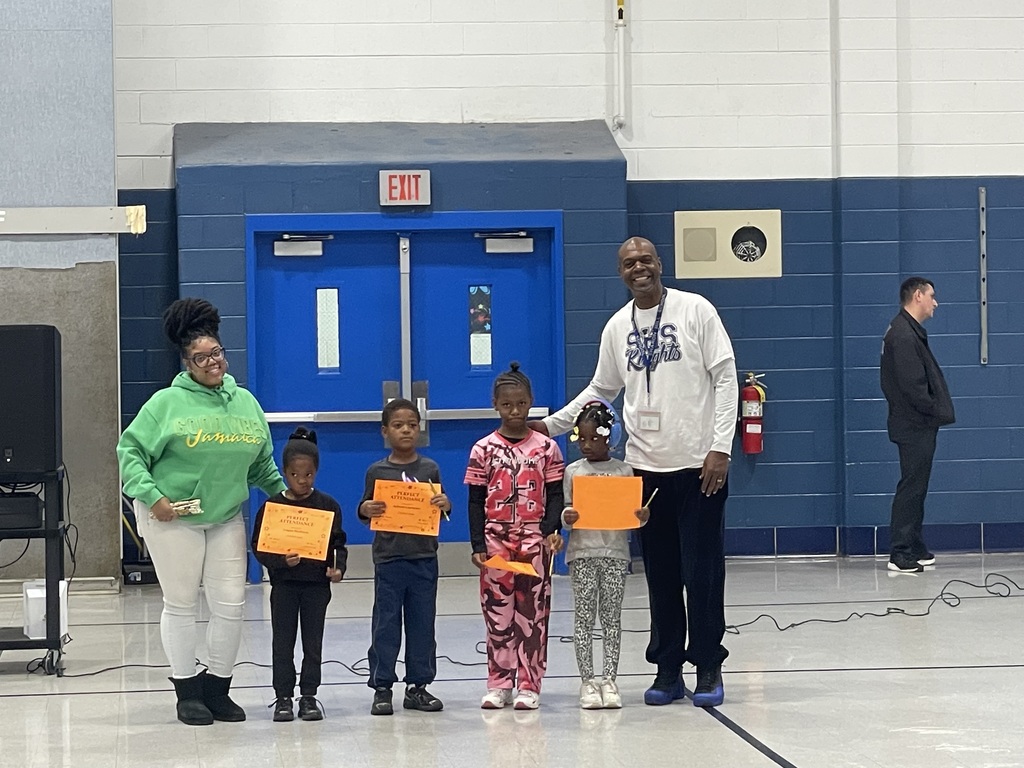 Four young students stand in a row holding orange award certificates, with two adults standing on either side of them in a school gym.