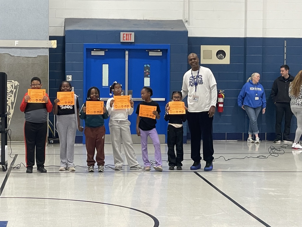 Six elementary students stand in a row holding orange award certificates in a school gym. An adult stands beside them smiling.