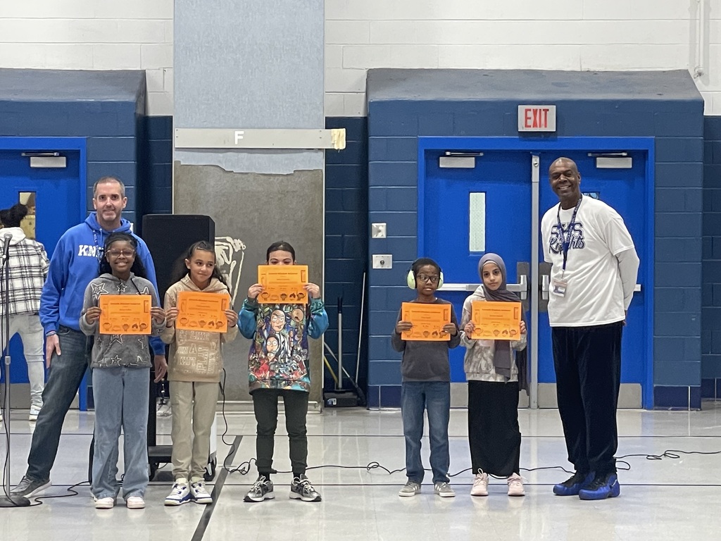 Six elementary students stand together in a gym holding orange award certificates, with an adult on each end of the group.