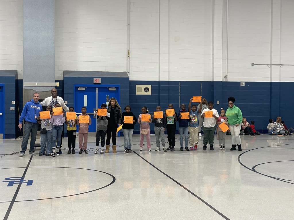 A long line of elementary students stand side by side in the gym holding orange award certificates. Two adults stand at each end of the line.