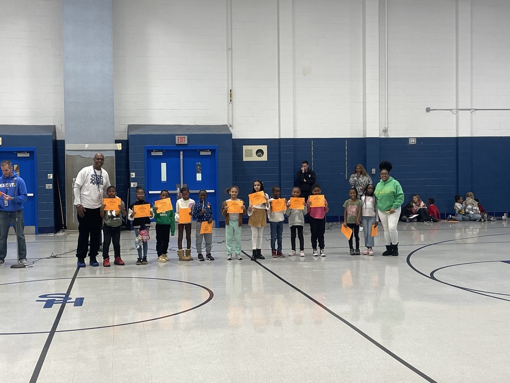 A group of young students stand in a row in the gym holding orange award certificates. An adult stands on each side of the group.