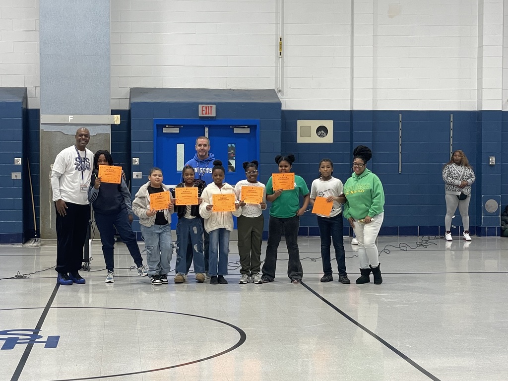 A group of elementary students stand in a gym holding bright orange award certificates. Two adults stand on each side of the group, smiling.