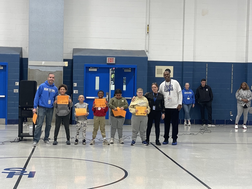 Seven students stand in a row in a gym, holding orange award certificates while two adults stand on either end. Several people are visible in the background near the wall.
