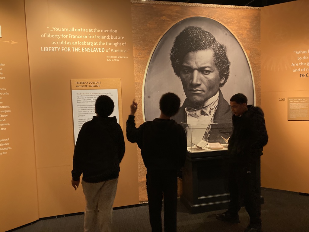 Three students stand in front of a museum exhibit featuring a large portrait of Frederick Douglass and text panels about his writings and advocacy.