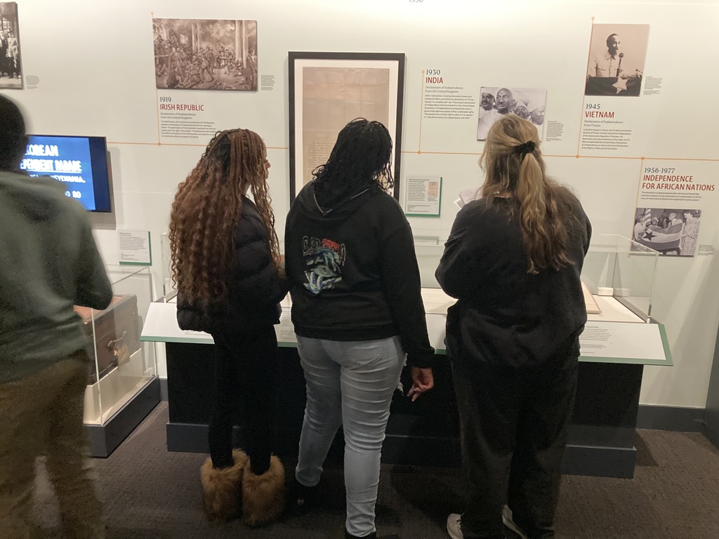 Three students stand closely together reading a museum display case with text and images about independence movements around the world.