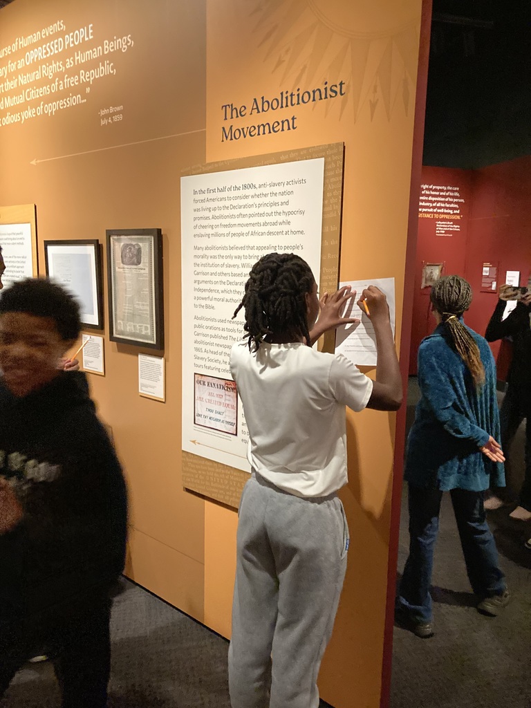 A student writes on a worksheet while standing in front of a museum panel titled “The Abolitionist Movement,” surrounded by other visitors.