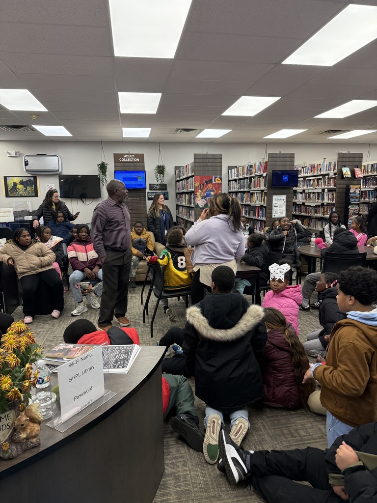 An adult speaks to a large group of seated children in a library, with students gathered on the floor and at tables, listening attentively.