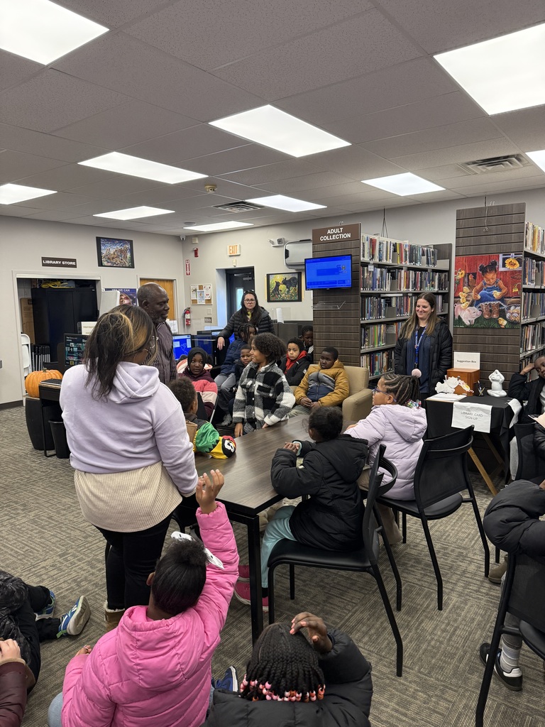 Elementary students sit around tables and on the floor in a library as an adult speaks to them