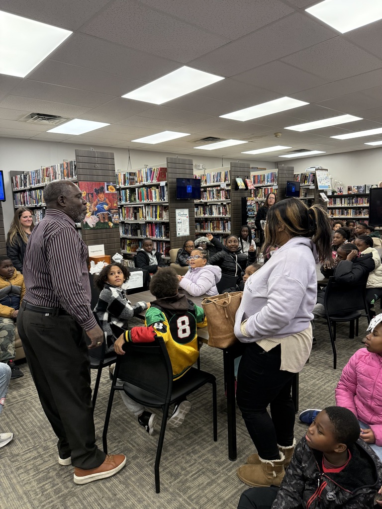 A speaker stands near a table addressing children seated on the floor and in chairs throughout a library
