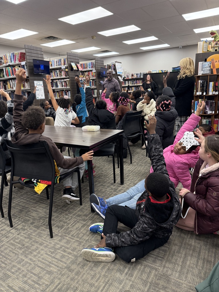 A large group of elementary students sit on the floor and at tables in a school library, raising their hands toward a man speaking at the front of the room while several adults stand nearby.