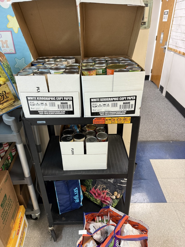 A black two-tier rolling cart loaded with food donation items. Two open copy-paper boxes filled with assorted canned goods sit on the top shelf.