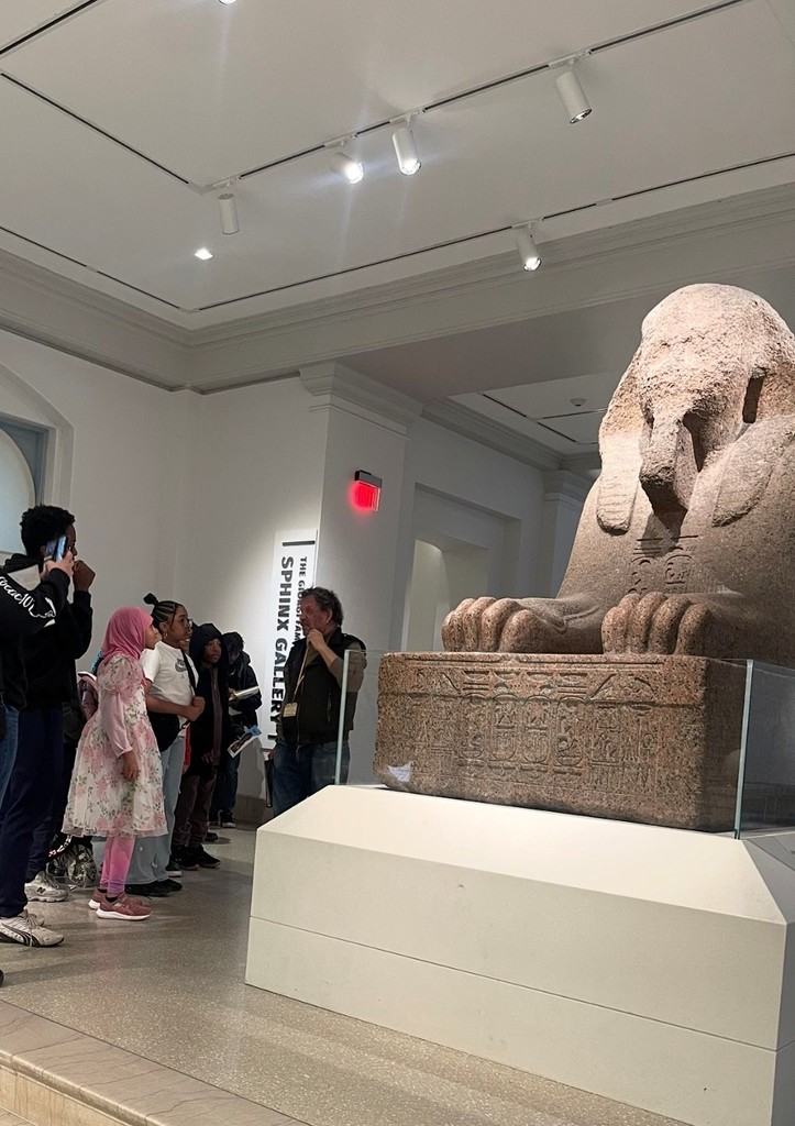 Students listen to a guide in the Sphinx Gallery while viewing a large ancient sphinx statue.