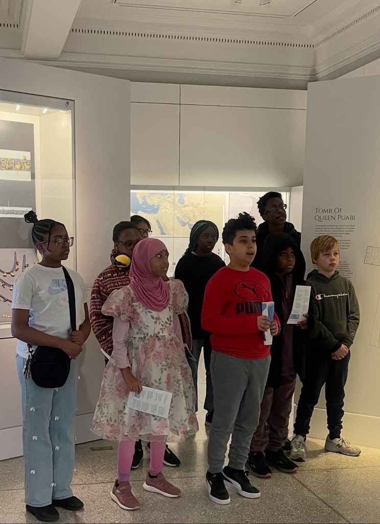 Students stand in a museum exhibit area labeled “Tomb of Queen Puabi,” holding pamphlets and listening.