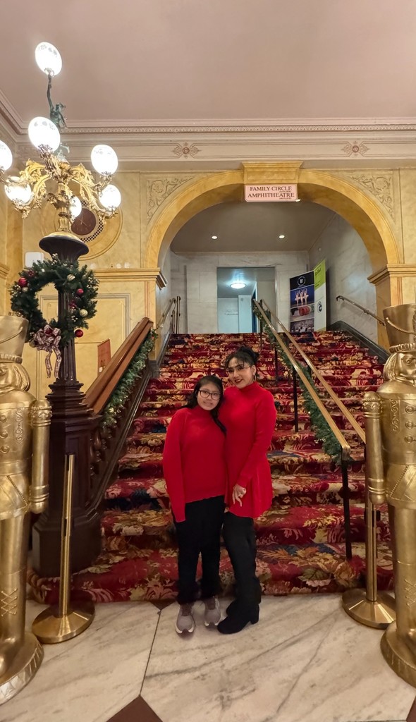 Two people in matching red outfits pose in front of a grand staircase decorated for the holidays at a theater.