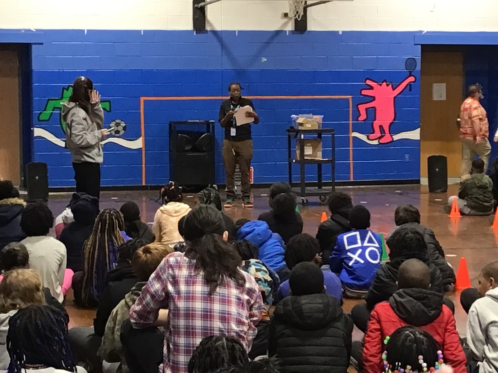 Children sit on the floor listening to a speaker holding papers in a school gym with bright blue walls and sports-themed artwork.