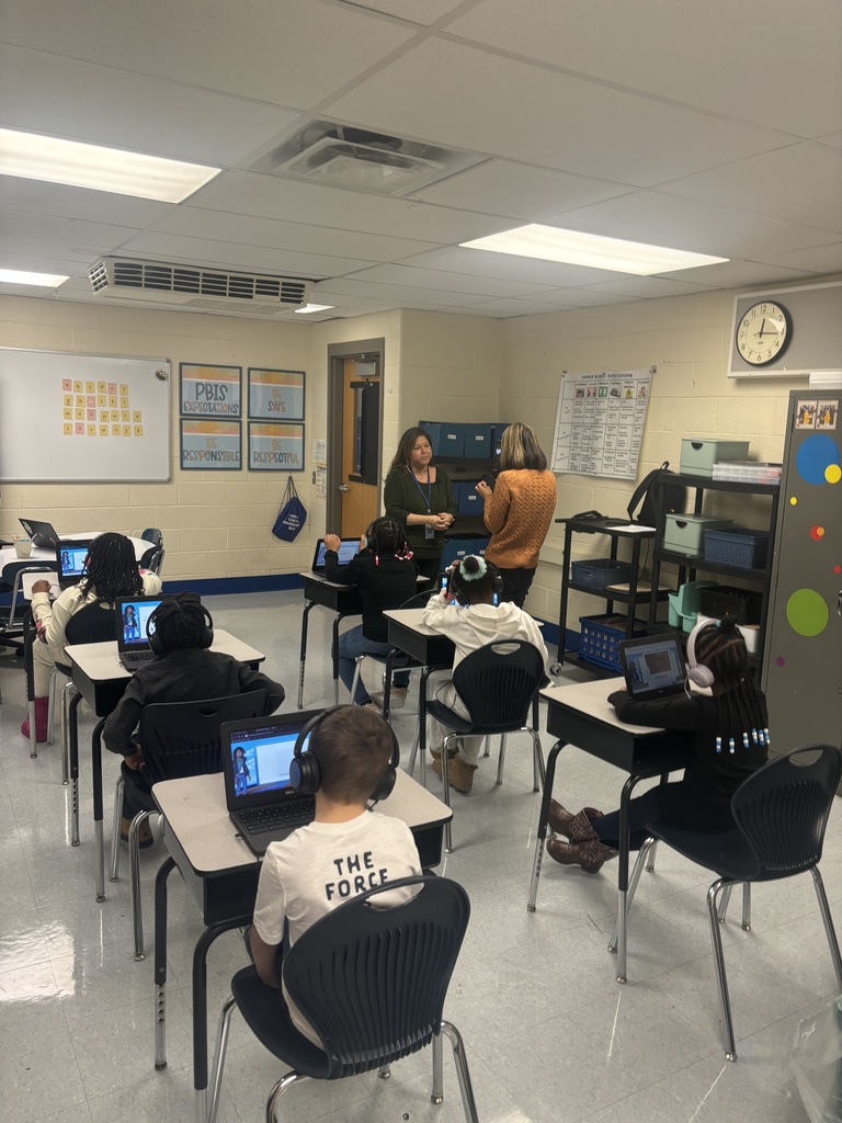 Elementary students wearing headphones and using laptops in a classroom while two teachers discuss near the whiteboard.