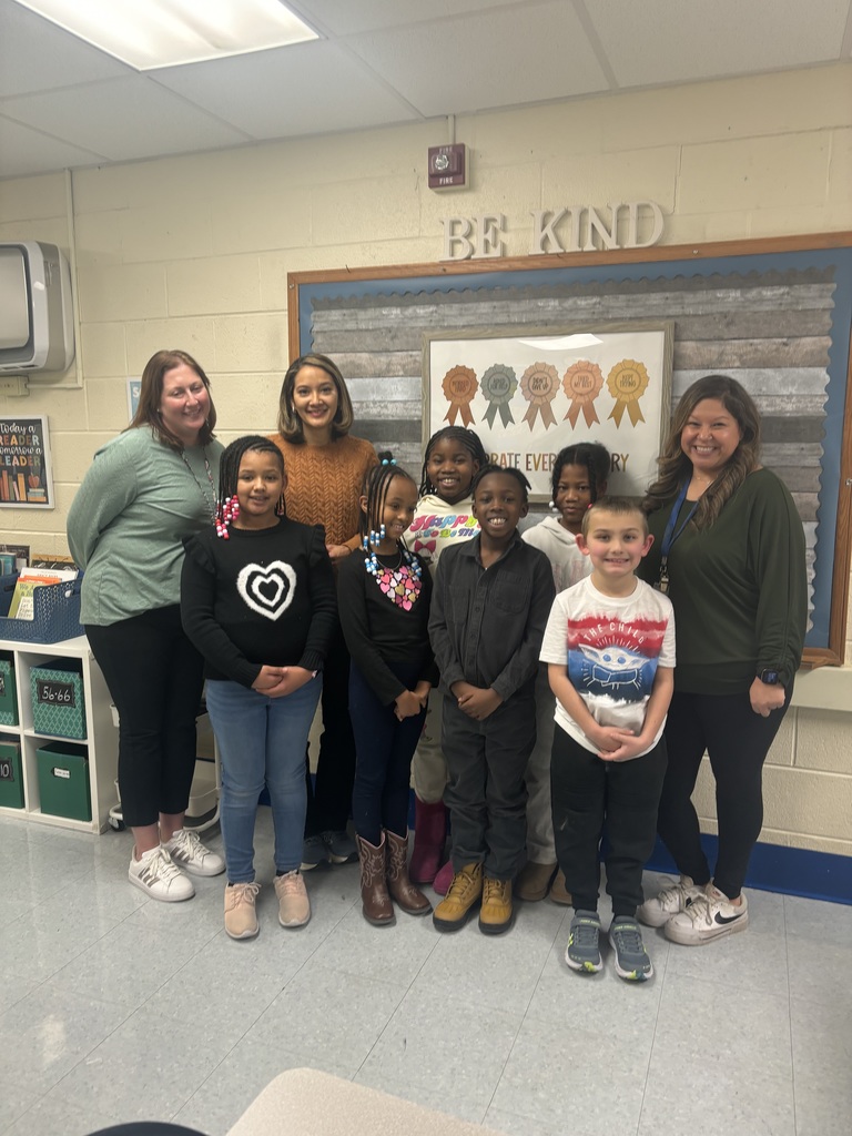 Group of children and adults standing together in a classroom with a 'Be Kind' sign on the wall behind them.