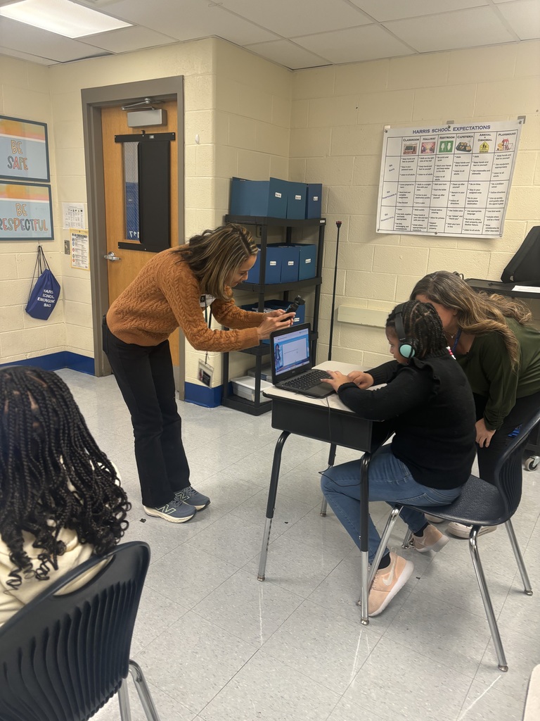 Teacher leaning over to take a photo of a student working on a laptop in a classroom setting.