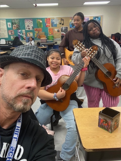 Three students in a classroom holding ukuleles, preparing to play music together during a lesson.