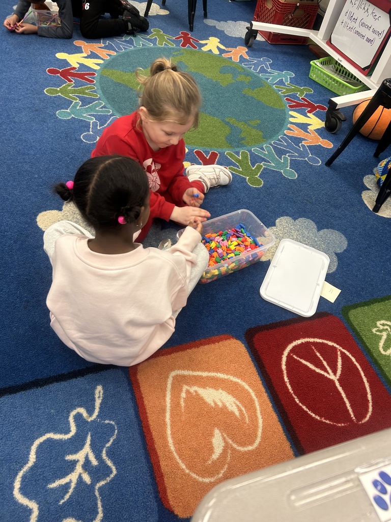 Two young children sitting on a colorful classroom carpet, playing with a container of small, colorful blocks together.