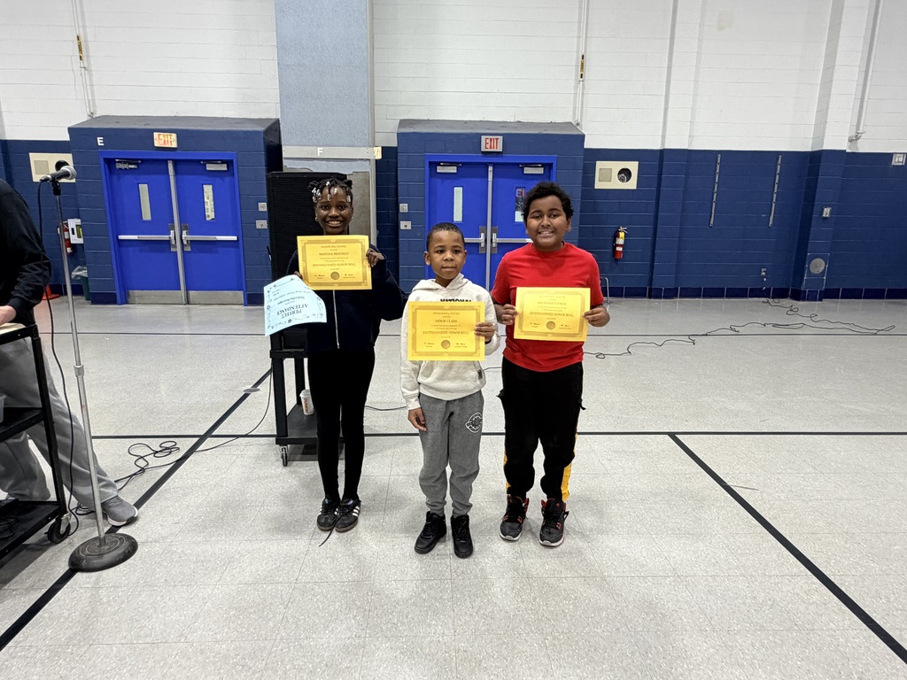 Three children standing in a gym holding yellow certificates, with blue doors and a microphone stand nearby.