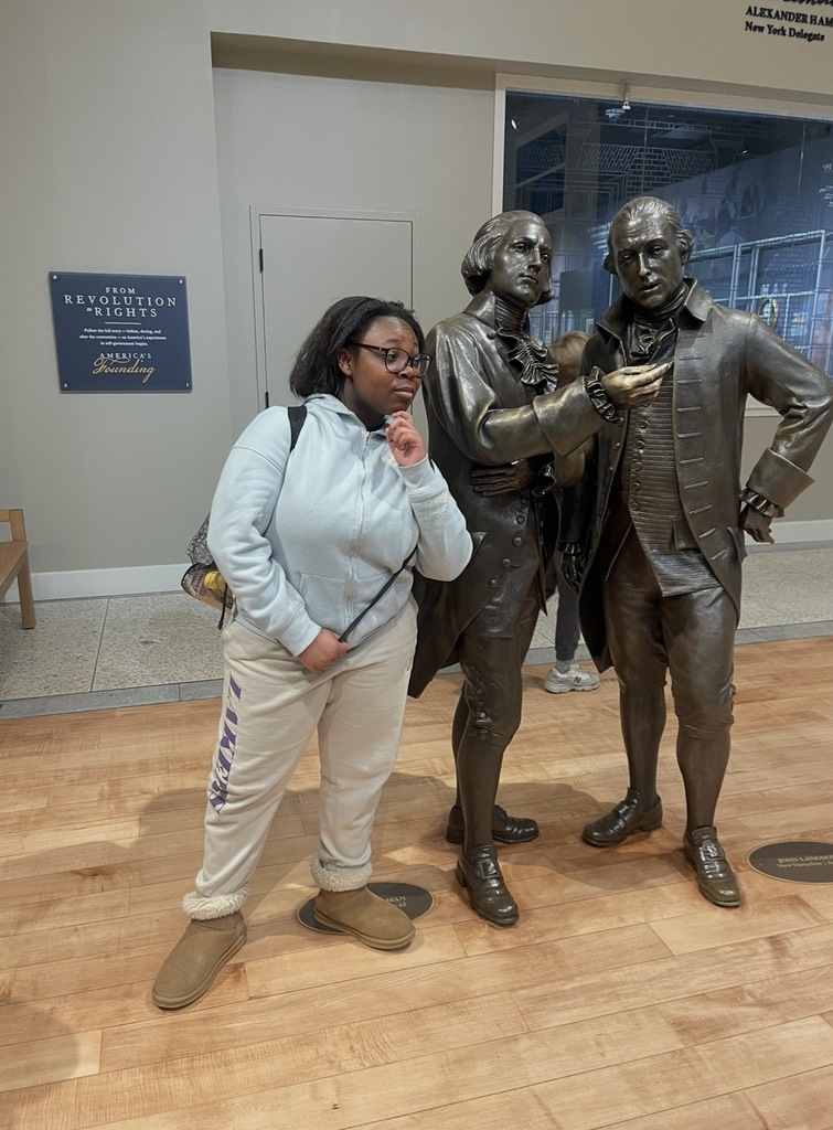 Person posing with two bronze statues of historical figures dressed in 18th-century attire inside a museum gallery.