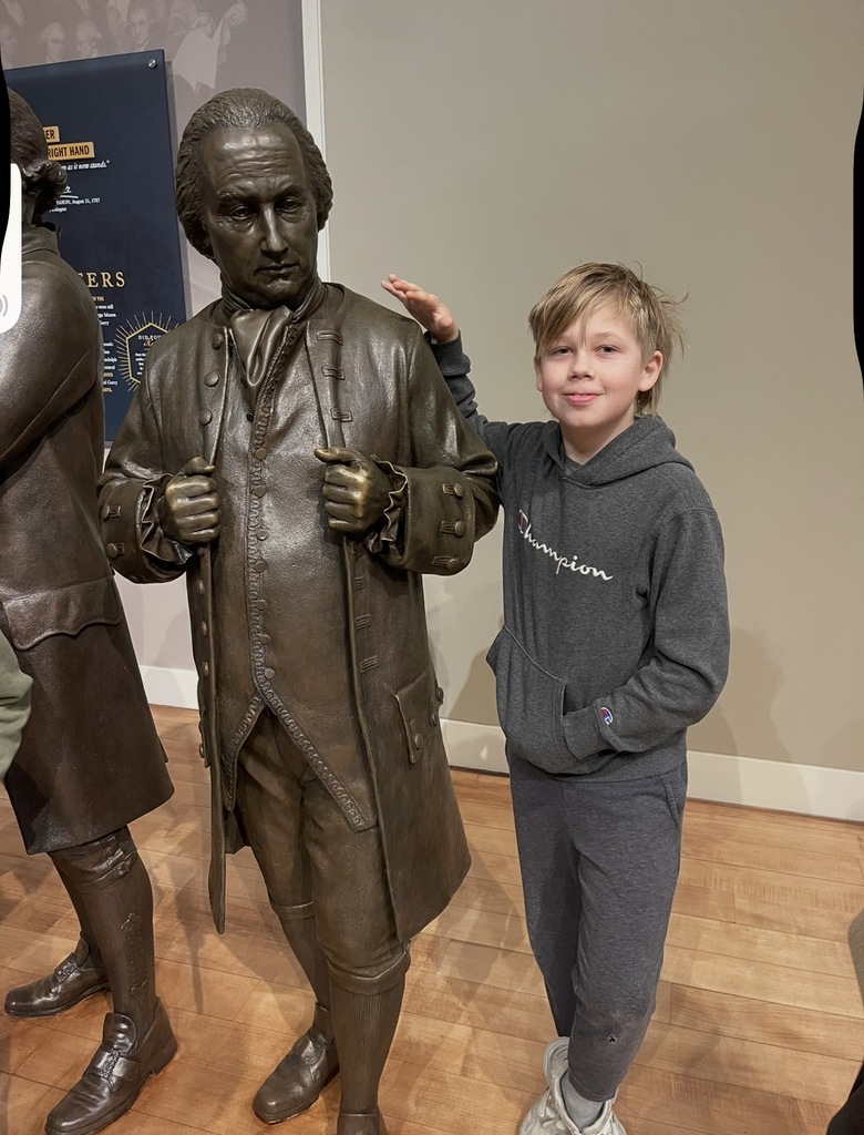 Young boy in gray hoodie posing next to a bronze historical statue inside a museum gallery.
