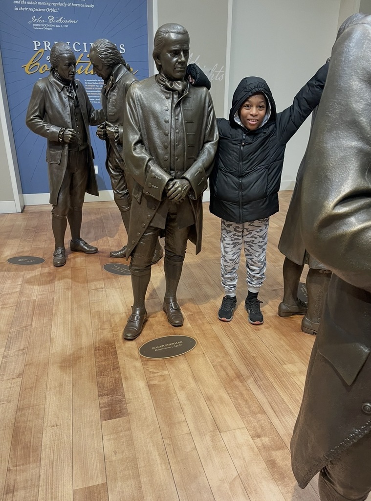 Child posing with life-sized bronze statues of historical figures dressed in 18th-century attire inside a museum gallery.
