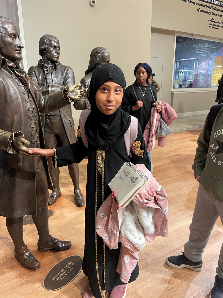Child wearing a black abaya and hijab stands inside a museum holding a pink jacket and a book, surrounded by bronze historical statues.