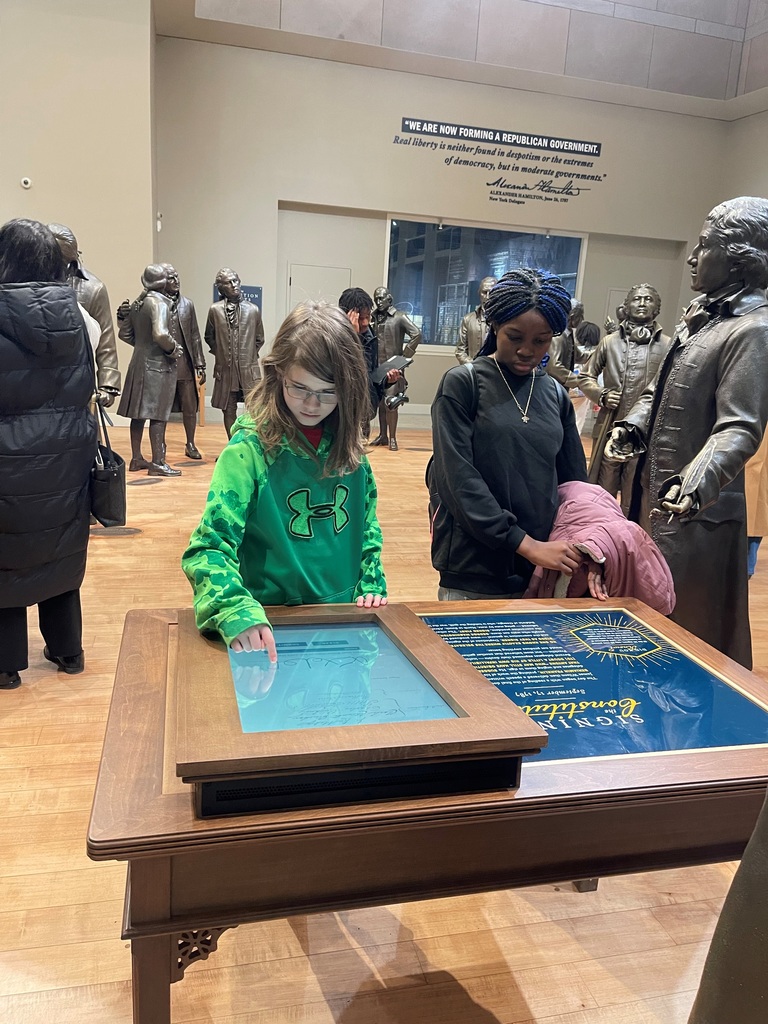 Children interacting with an educational touchscreen table in a museum surrounded by bronze statues of historical figures.