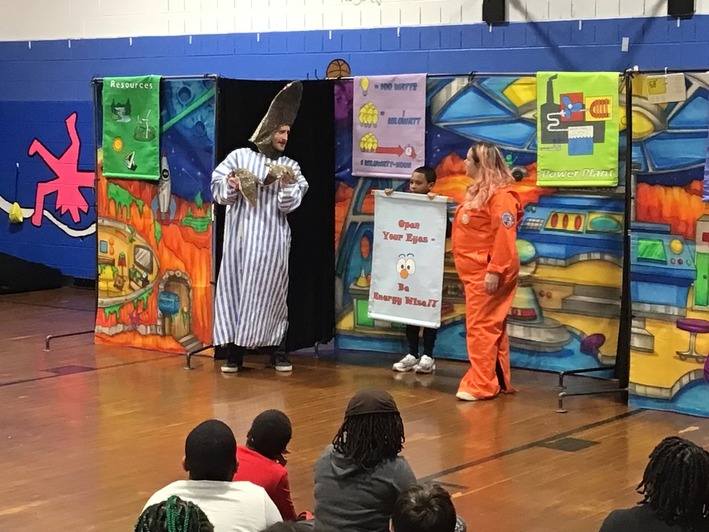 Actors in colorful costumes perform an educational play about energy conservation for children in a school gymnasium.