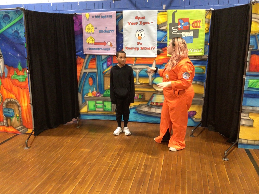 Person in an orange astronaut suit holding a model of the solar system while talking to a child in black clothing inside a colorful educational space exhibit.