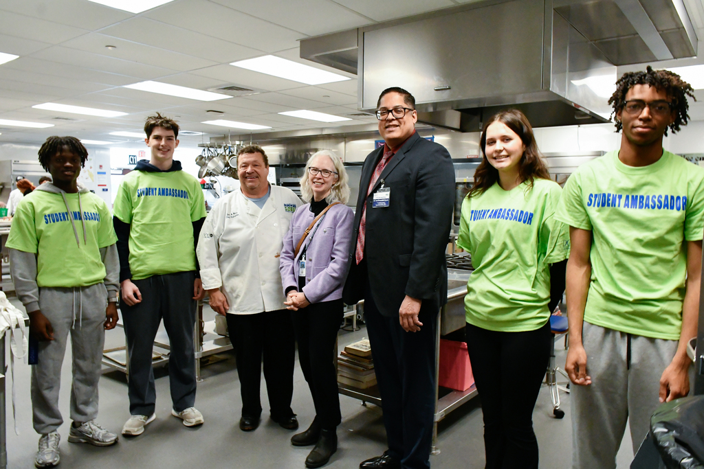 Group of students wearing green Student Ambassador shirts standing with two adults in a professional kitchen setting.