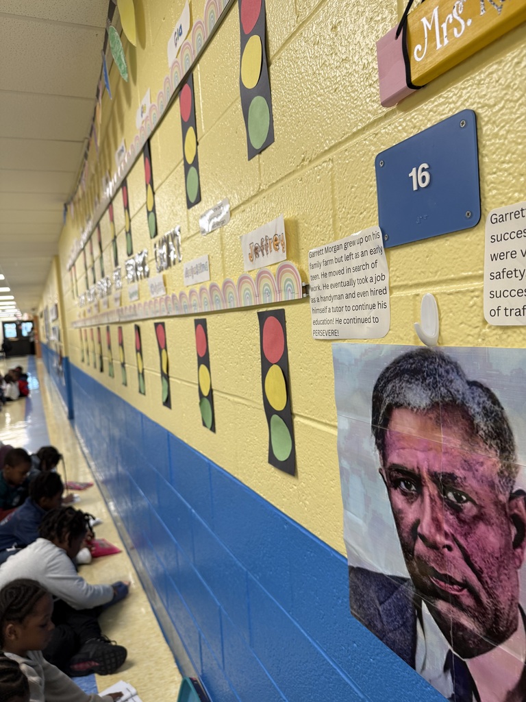 Elementary students sitting along a school hallway wall decorated with colorful traffic light cutouts and educational posters.
