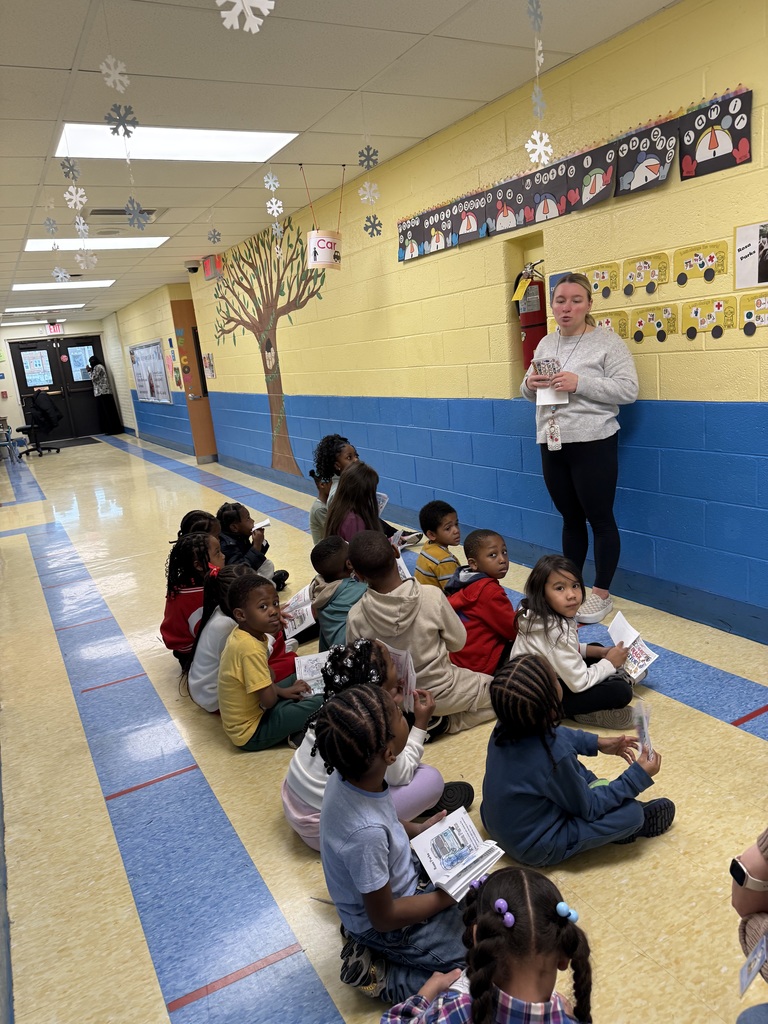 A group of young students sitting on a school hallway floor listening to a teacher standing and holding papers.