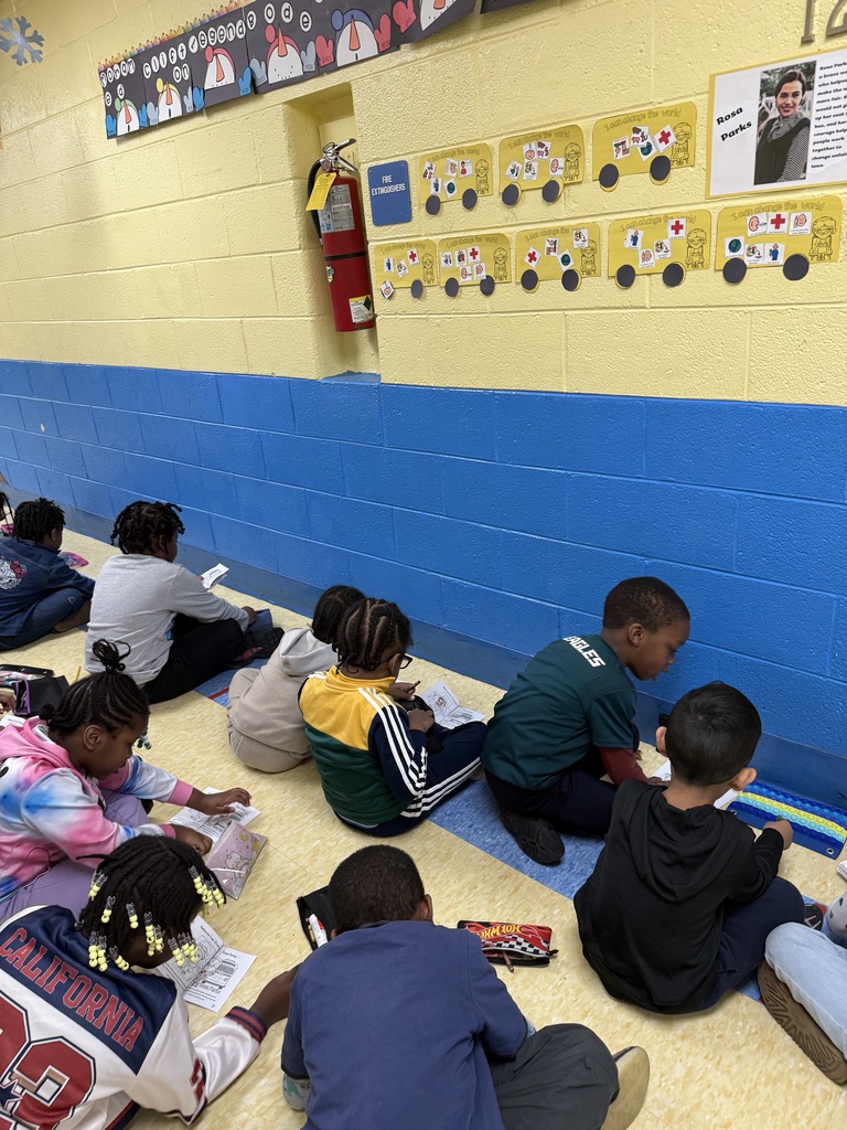 A group of young students sitting on the floor in a classroom, engaged in reading or writing activities near a blue and yellow painted wall.
