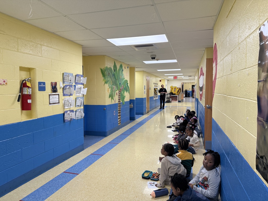 Children sitting on the floor along a school hallway with yellow and blue walls, listening to a teacher in the distance.