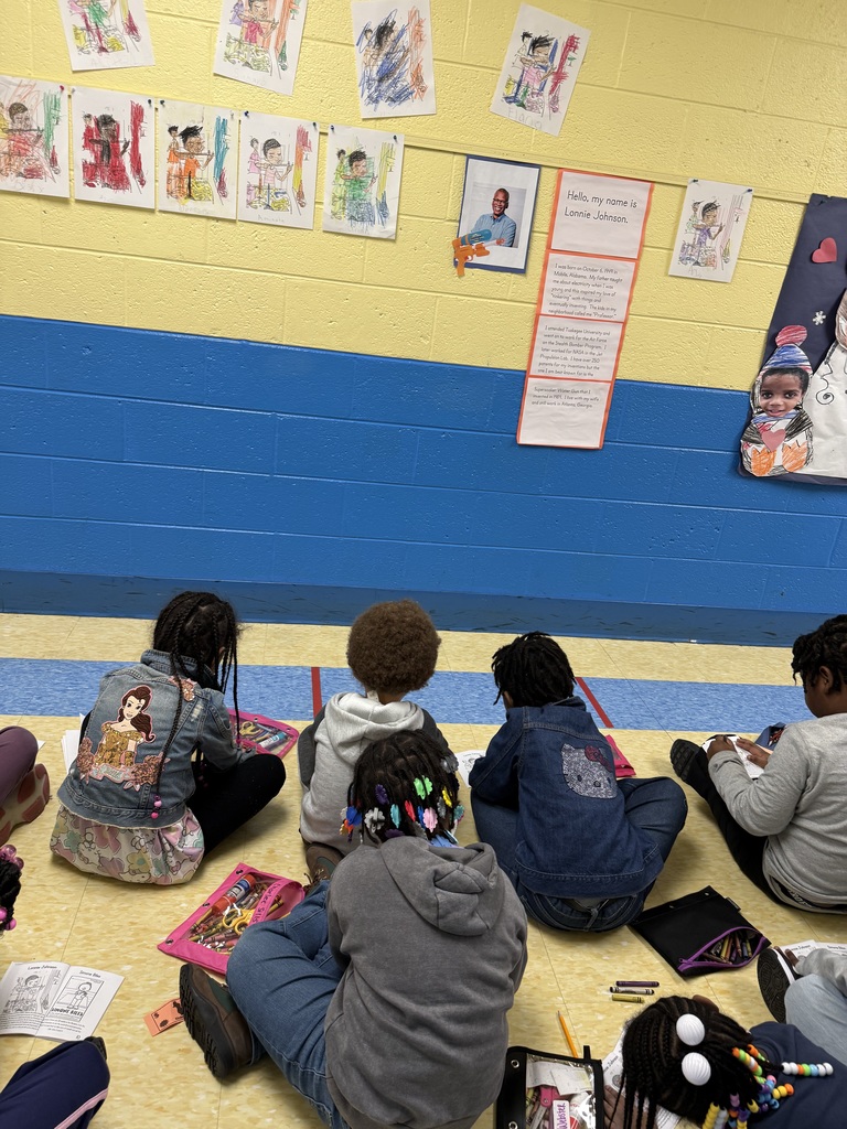 Children sitting on the floor along a school hallway with yellow and blue walls, listening to a teacher in the distance.