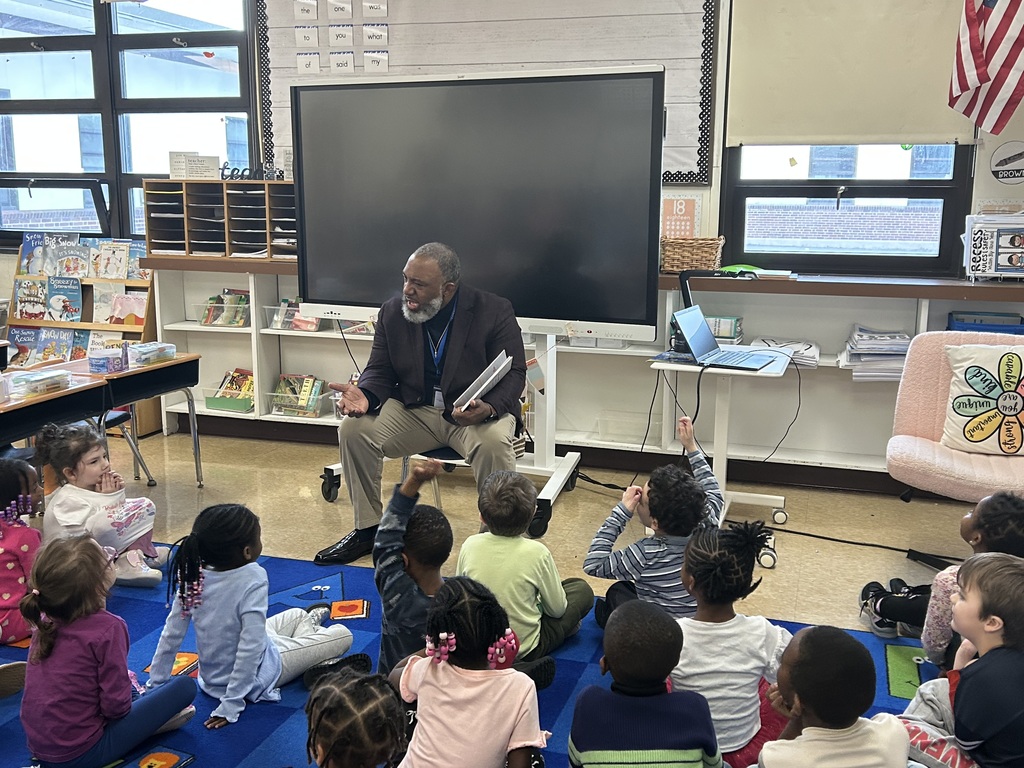 Teacher reading a book to a diverse group of young children seated on a colorful classroom rug.